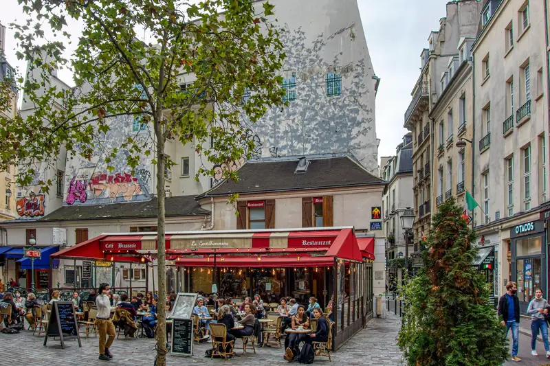 Café-brasserie with red awnings and a busy terrace on Place Saint-André-des-Arts in Paris, with a Metro sign on the left