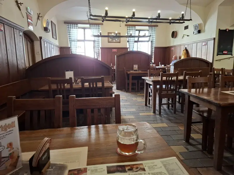 A glass mug of semi-dark beer on a coaster on a wooden table in the U Medvídků pub in Prague, with empty wooden booths and chairs behind
